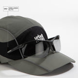 Side view of olive-grey and black color-block nylon cap with white stacked logo and pine tree icon and sunglasses slits. Urban Monkey®