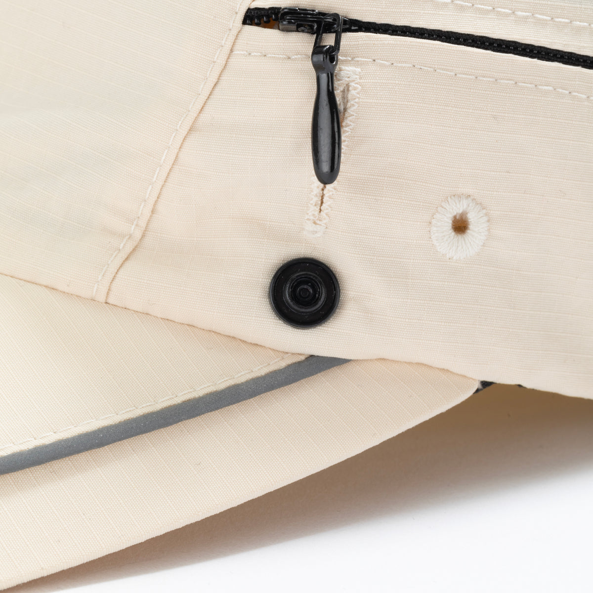 Close-up view of cream ripstop nylon cap showing black zipper pull, reinforced slit, snap button, eyelet vent and reflective brim piping. Urban Monkey®