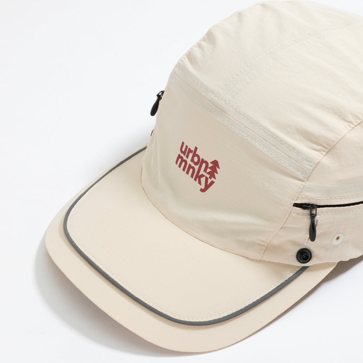 Front top view of cream nylon sun cap with rust-red embroidered logo, zip pocket, and snap button. Urban Monkey®